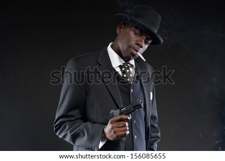 Retro african american mafia man wearing striped suit and tie and black hat. Holding a gun. Smoking cigarette. Studio shot.