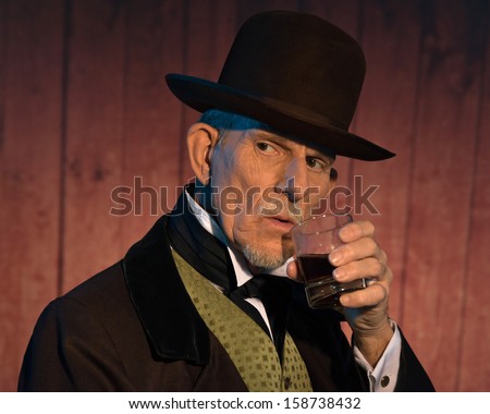 Senior alert western man wearing a brown hat and coat holding a whiskey. In front of wooden wall in saloon.