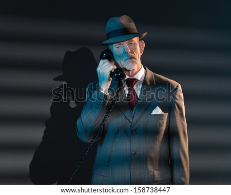 Retro detective man calling with vintage telephone at night in office. Lit by light through venetian blinds.