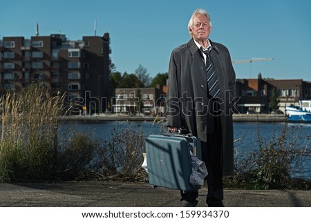 Senior business man without a job and homeless on the street. Holding a suitcase. Dirty suit and raincoat.