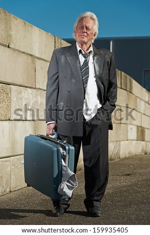 Depressed senior business man without a job and homeless on the street. Holding a suitcase. Wearing a dirty suit.