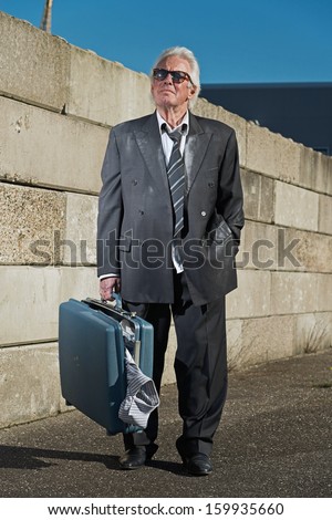 Depressed senior business man with sunglasses without a job and homeless on the street. Holding a suitcase. Wearing a dirty suit.