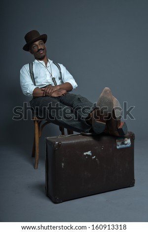Retro afro america western cowboy man with mustache. Sitting in wooden chair. Resting his legs on suitcase. Wearing brown hat. Cool tough guy.