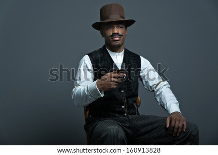 Vintage afro america western cowboy man with mustache. Drinking whiskey. Sitting in wooden chair. Wearing brown hat. Cool tough guy.