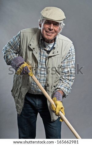 Smiling senior gardener man with hat holding hoe. Studio shot against grey.
