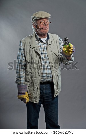 Gardener senior man with hat holding pruning shears. Wearing glasses and smoking pipe. Studio shot against grey.