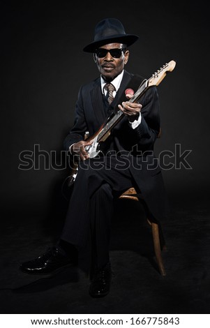 Retro senior afro american blues man. Wearing striped suit with blue hat and black sunglasses. Playing electric guitar.