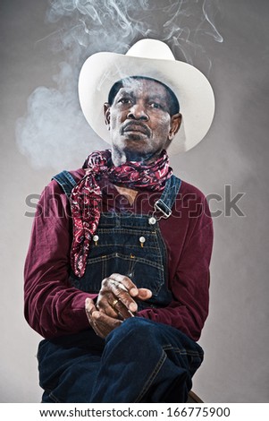 Retro senior afro american blues man in times of slavery. Wearing denim bib and brace overall with white hat. Smoking a cigarette.