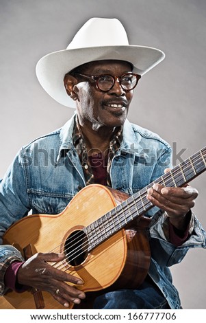 Retro senior afro american blues man in times of slavery. Wearing jeans jacket with white hat and glasses. Playing acoustic guitar.