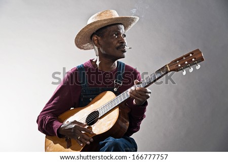 Retro senior afro american blues man in times of slavery. Wearing denim bib and brace overall with straw hat. Playing acoustic guitar.