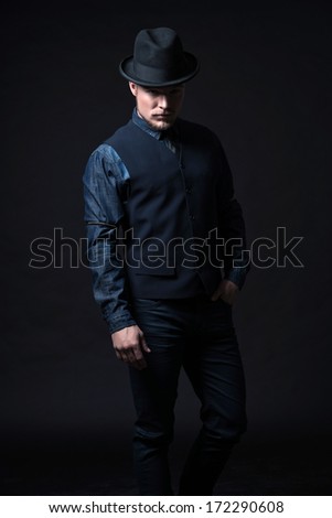 Retro 1900 modern fashion man. Wearing blue jeans shirt with gilet and trousers. Black hat. Studio shot against black.