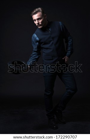 Retro 1900 modern fashion man. Wearing blue jeans shirt with gilet and trousers. Holding black hat. Studio shot against black.