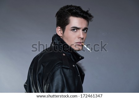 Retro rock and roll 50s fashion man with dark grease hair. Smoking cigarette. Wearing black leather jacket. Studio shot against grey.
