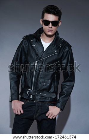 Macho retro rock and roll fifties fashion man with dark grease hair. Wearing black leather jacket and sunglasses. Studio shot against grey.