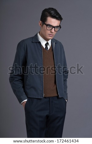 Retro Fifties Fashion Man With Dark Grease Hair. Wearing Brown Sweater With Blue Jacket And Black Glasses. Studio Shot Against Grey.