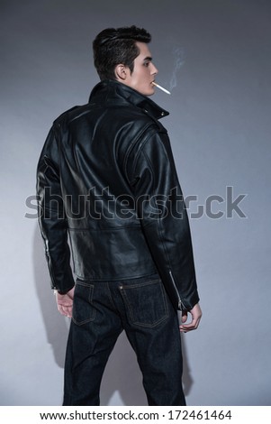 Retro rock and roll 50s fashion man with dark grease hair. Smoking cigarette. Wearing black leather jacket. Studio shot against grey.
