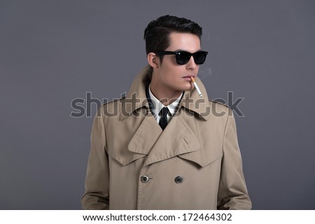 Retro fifties business fashion man with dark grease hair. Wearing brown raincoat and black sunglasses. Smoking cigarette. Studio shot against grey.