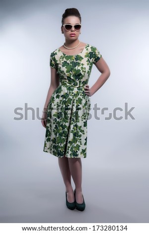 Retro 50s fashion brunette girl with sunglasses wearing green dress and white pearl necklace. Studio shot against grey.