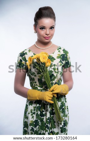 Retro 50s fashion housewife wearing yellow rubber gloves. Holding flowers. Studio shot against grey.
