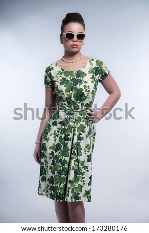 Retro 50s fashion brunette girl with sunglasses wearing green dress and white pearl necklace. Studio shot against grey.