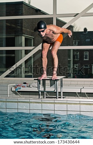 Healthy active senior man with beard in indoor swimming pool diving from starting block. Wearing swim cap and glasses.