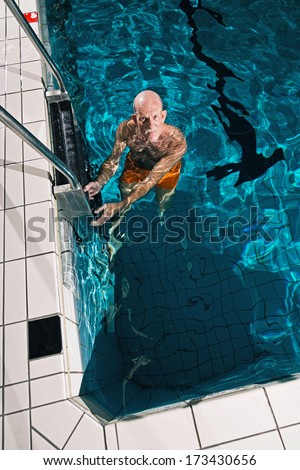Healthy active senior man with beard in indoor swimming pool. Wearing orange swimming trunks.