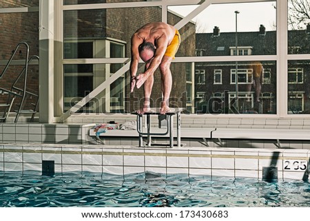 Healthy active senior man with beard in indoor swimming pool diving from starting block. Wearing yellow swimming trunks.