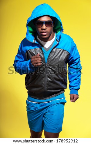 Athletic black man in sportswear fashion. Runner with hoody jacket and sunglasses. Intense colors. Studio shot against yellow.