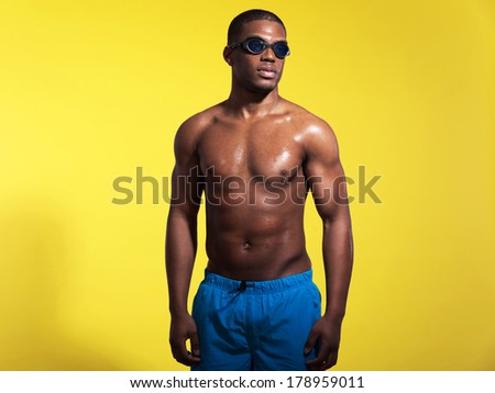 Athletic swimmer with waterdrops. Black man wearing blue swimming shorts and protective glasses. Intense colors. Studio shot against yellow.