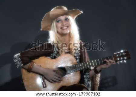 Retro soft focus hippie 70s country guitar girl with long blonde hair. Wearing a hat. Studio shot against grey.