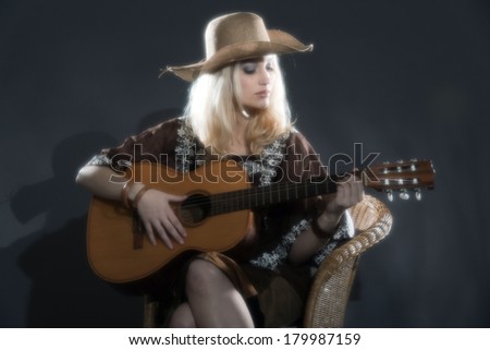 Retro soft focus hippie 70s country guitar girl with long blonde hair. Wearing a hat. Studio shot against grey.