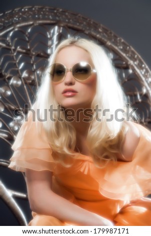 Retro hippie 70s fashion sensual girl with long blonde hair wearing an orange dress and sunglasses. Sitting on a big chair. Studio shot against grey.
