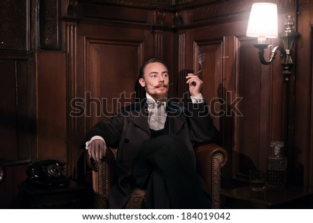 Vintage 1900 fashion man with beard. Sitting in old wooden reading room. Smoking a cigar.