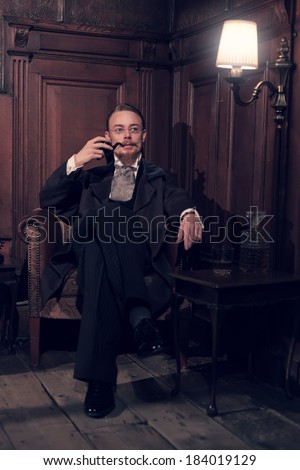 Vintage 1900 fashion man with beard and glasses. Sitting in old wooden reading room. Smoking pipe.