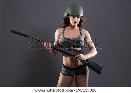Muscled fitness army girl holding gun. Wearing green helmet. Studio shot against grey.