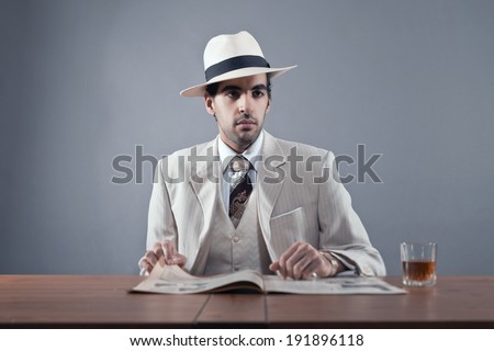 Mafia fashion man wearing white striped suit and hat. Sitting at table with glass of whisky and newspaper. Studio shot.