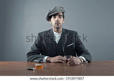 Mafia fashion man wearing grey striped suit with cap. Sitting at table with glass of whisky and cigarettes. Studio shot.