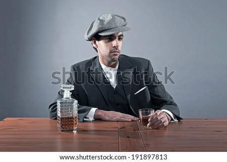 Mafia fashion man wearing grey striped suit with cap. Sitting at table with glass of whisky and cigarettes. Studio shot.
