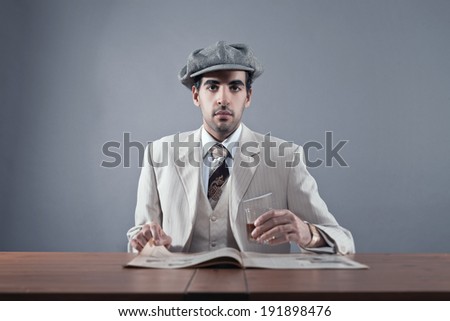 Mafia fashion man wearing white striped suit and cap. Sitting at table with glass of whisky and newspaper. Studio shot.