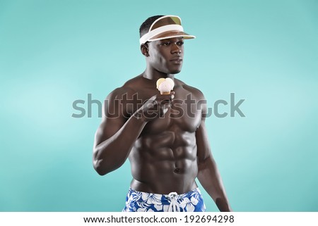 Summer black african american muscled fitness man holding ice cream. Wearing blue swimming shorts and cap. Studio shot against blue.