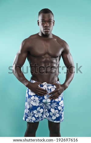 Black african american muscled fitness man. Summer swimwear fashion. Wearing blue swimming shorts. Studio shot against blue.