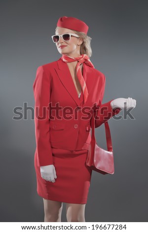 Retro blonde stewardess wearing red suit with cap and sunglasses. Holding red bag. Studio shot against grey.