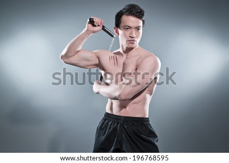 Muscled asian kung fu man in action pose with nunchucks. Blood stripes on his chest. Wearing black pants. Studio shot against grey.