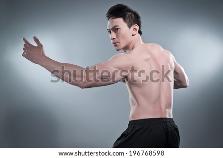 Muscled asian kung fu man in action pose. Wearing black pants. Studio shot against grey.