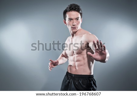 Muscled asian kung fu man in action pose. Blood stripes on his chest and face. Wearing black pants. Studio shot against grey.