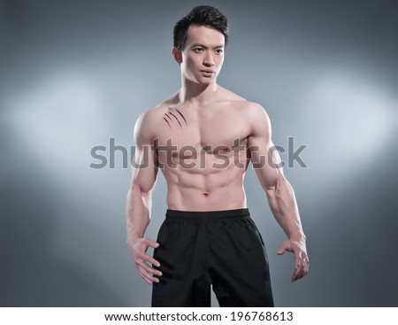 Muscled asian kung fu man in action pose. Blood stripes on his chest. Wearing black pants. Studio shot against grey.