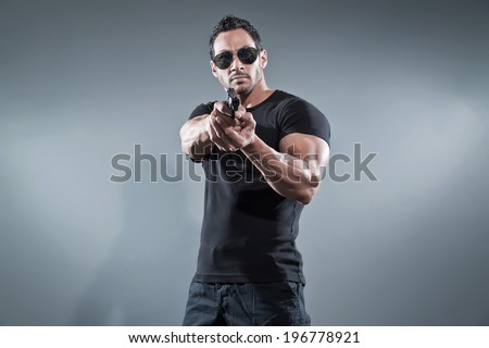 Action hero muscled man shooting with gun. Wearing black t-shirt with pants and sunglasses. Studio shot against grey.