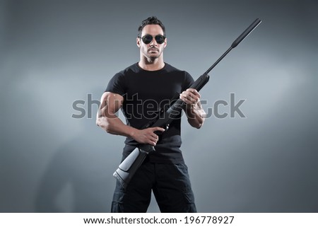 Action hero muscled man holding a rifle. Wearing black t-shirt with pants and sunglasses. Studio shot against grey.