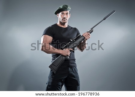 Commander muscled action hero man with rifle wearing black t-shirt and pants. Studio shot against grey.