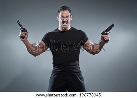 Action hero muscled man holding two guns. Wearing black t-shirt and pants. Studio shot against grey.